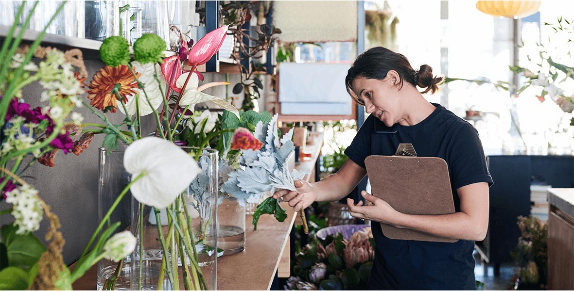 A woman on the phone working in a flower shop looking at a variety of plants.
